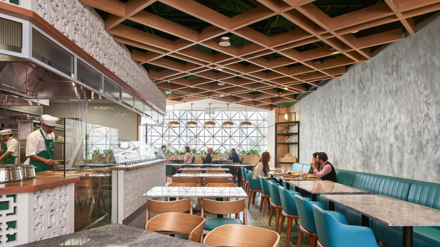Gensler: Restaurant Interior Design Inspiration. A wide restaurant room, with four tables with sofa seating on one of the walls, and many other dining tablesfor four people. You can see the kitchen through the window.